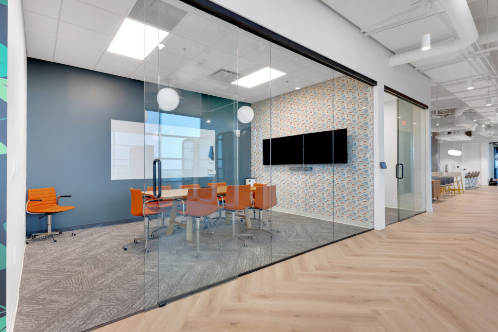 A glass-walled conference room with a table, six orange chairs, a whiteboard, and a wall-mounted monitor against patterned wallpaper.