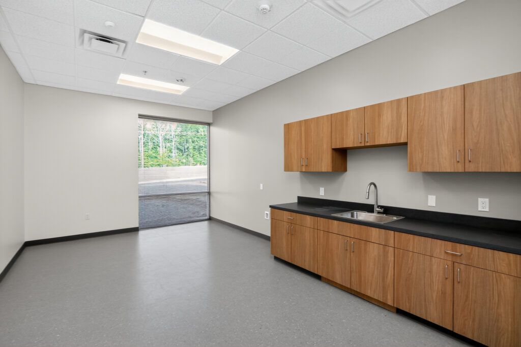An empty office breakroom with wood-finish cabinets, a black countertop with a stainless steel sink, and a large window overlooking trees.