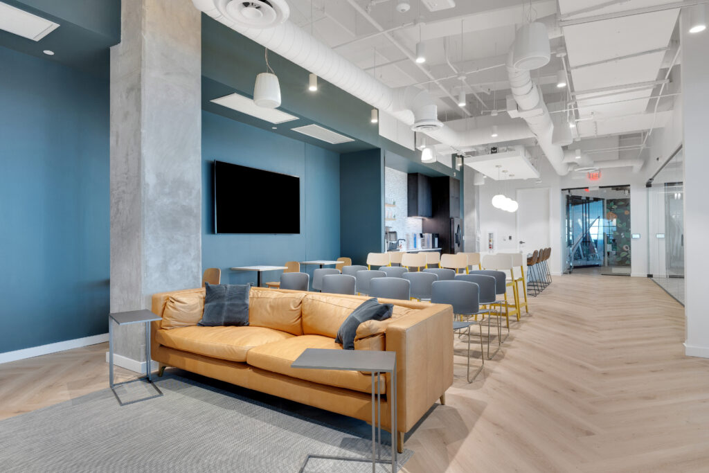 Office lounge area featuring a tan leather sofa, rows of chairs, and a kitchenette in the background.