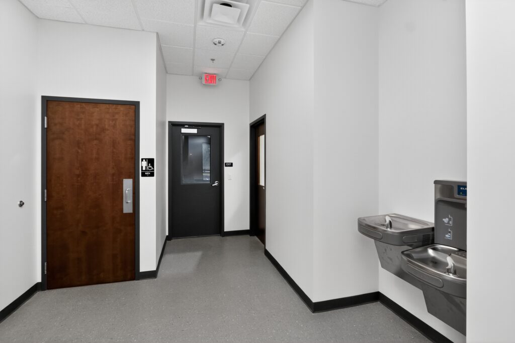A hallway featuring a wooden men’s restroom door, a door labeled exit, and a dual-level water fountain.