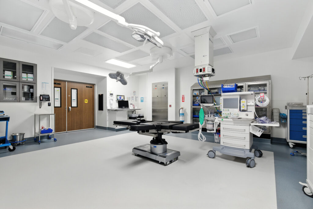 An operating room with a surgical table in the center, surrounded by medical equipment, a monitor, and overhead surgical lights.