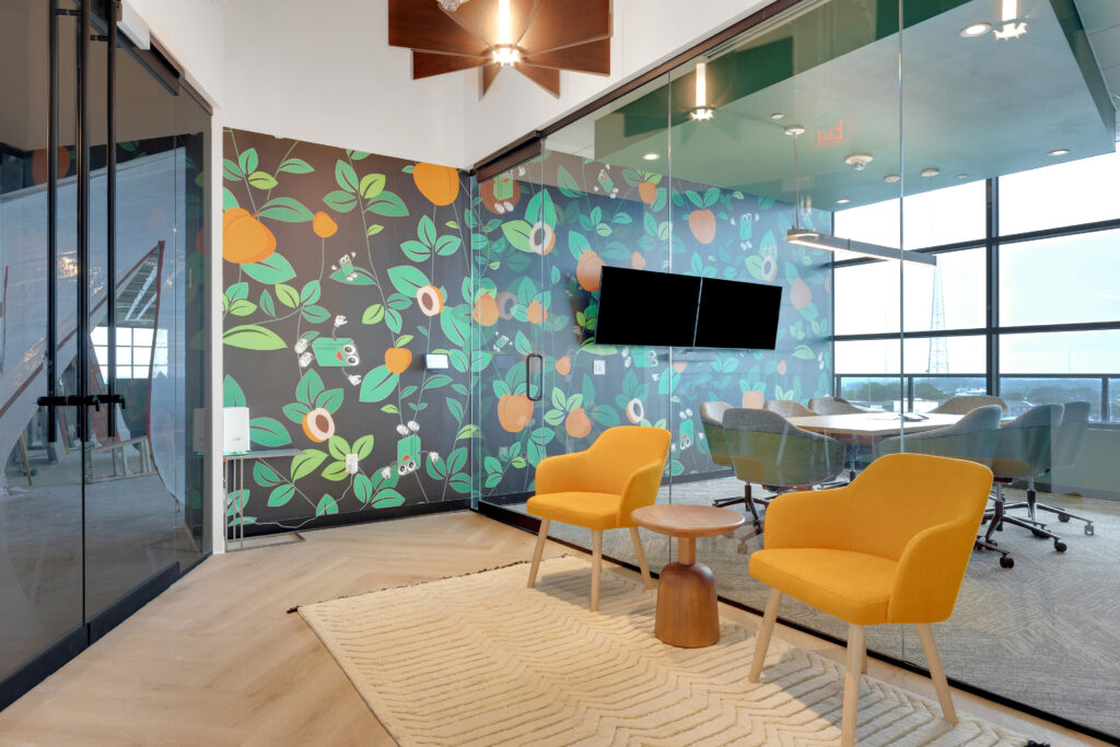 Two yellow armchairs and a small wooden table on a textured rug in front of a glass-walled conference room with floral wallpaper and a flat-screen television.