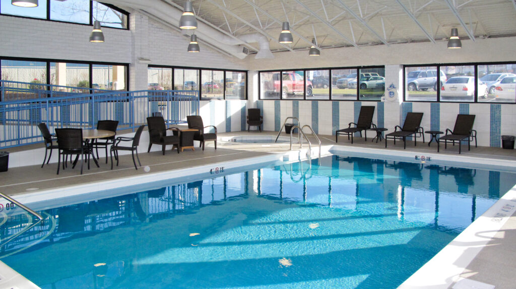 Indoor swimming pool with lounge chairs, tables, and a small hot tub, surrounded by large windows.