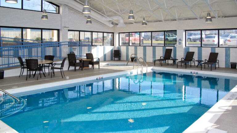 Indoor swimming pool with lounge chairs, tables, and a small hot tub, surrounded by large windows.