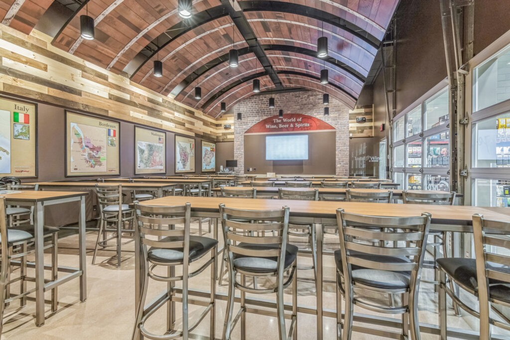 Interior of a wine shop with rows of high-top tables and chairs, a curved wooden ceiling, and maps of wine regions displayed on the walls.