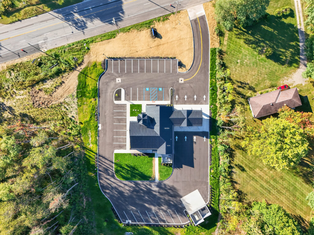Aerial view of a commercial building with a parking lot and drive-thru lane, situated between a road and wooded area.