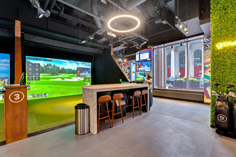 Indoor golf simulator bay with a large digital screen, high-top seating, and a golf bag standing against a green wall.