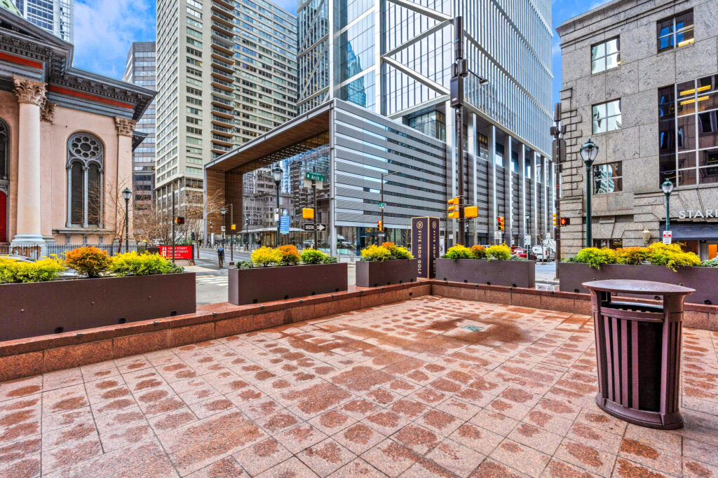 A paved outdoor plaza with planters in front of modern skyscrapers and a historic stone building at a city intersection.