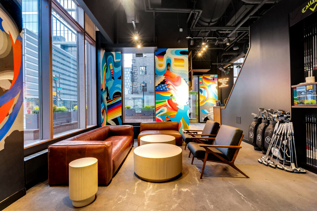 Lounge area with a leather sofa, two armchairs, and two cylindrical coffee tables, set against walls with vibrant abstract murals.