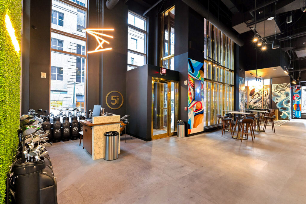 Indoor golf facility with a reception desk, golf bag storage, and high-top seating near floor-to-ceiling windows.
