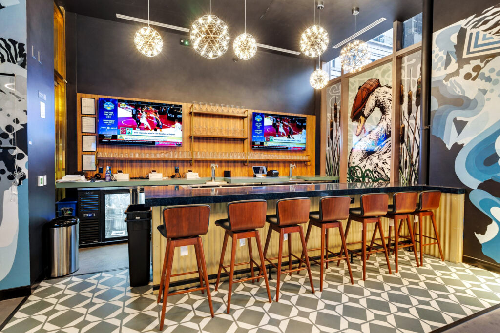 A modern bar with six wooden stools at a counter, two televisions showing basketball games, and a large wall mural of a swan.