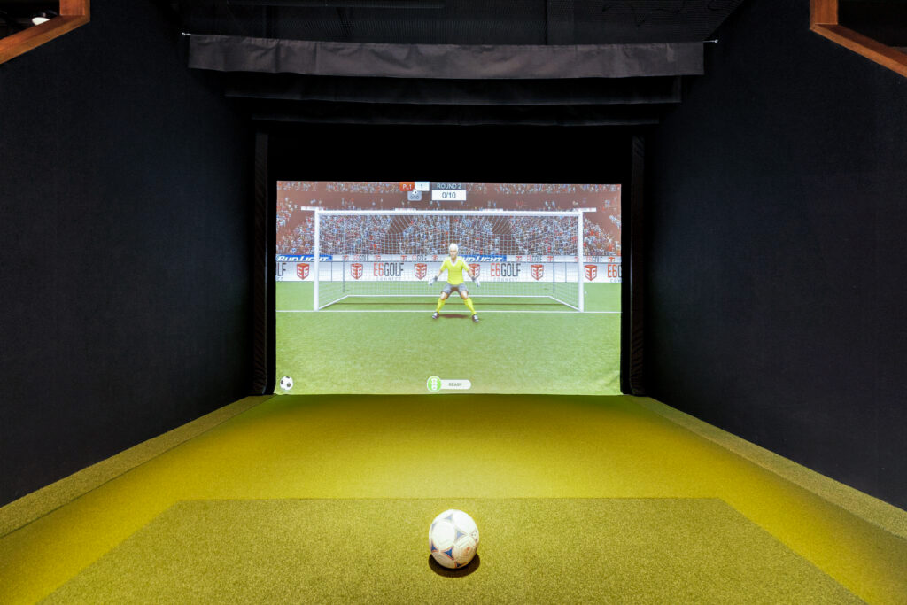 A soccer ball on green turf facing a large simulator screen displaying a goalkeeper in a stadium.