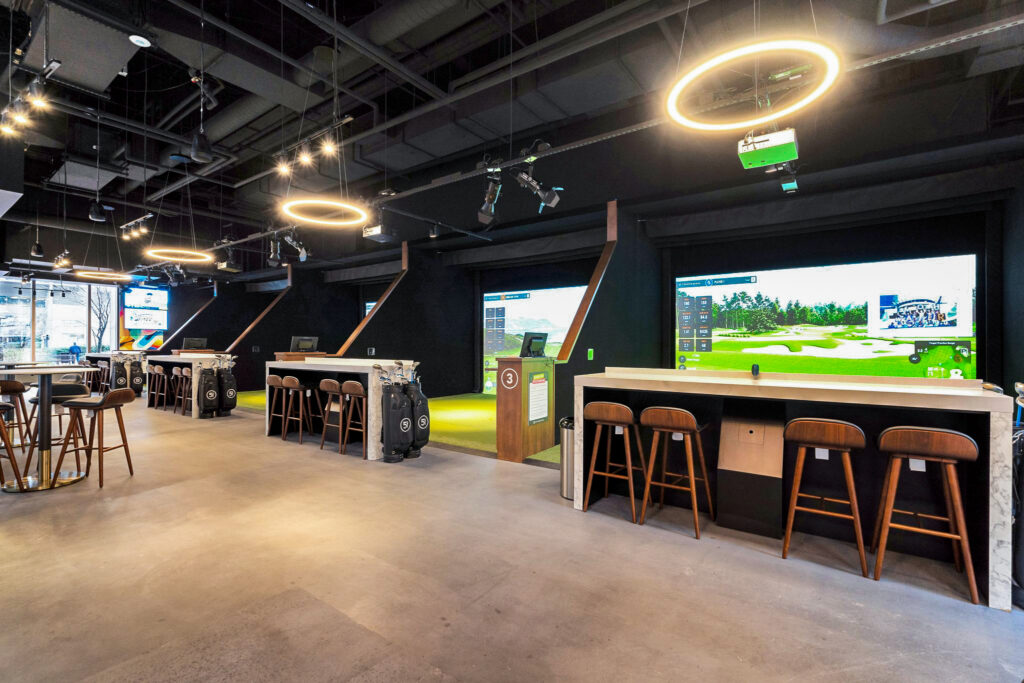 Indoor golf simulator bays with high-top tables, bar stools, and digital screens displaying golf courses.