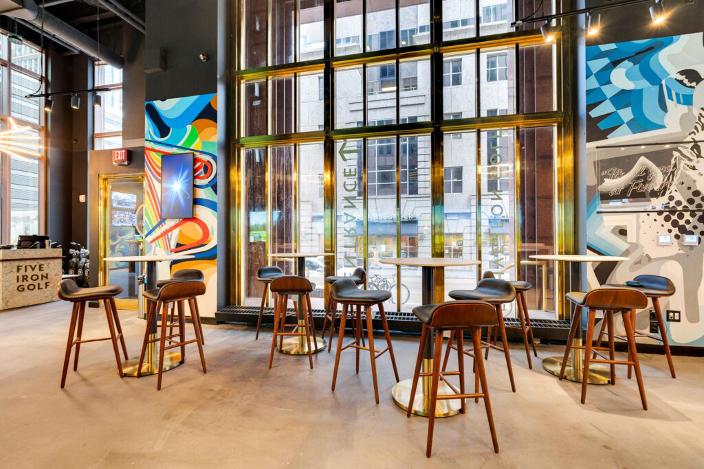 Modern lounge with tall stools at high-top tables, large windows, and colorful mural walls, with a “Five Iron Golf” sign in the corner.