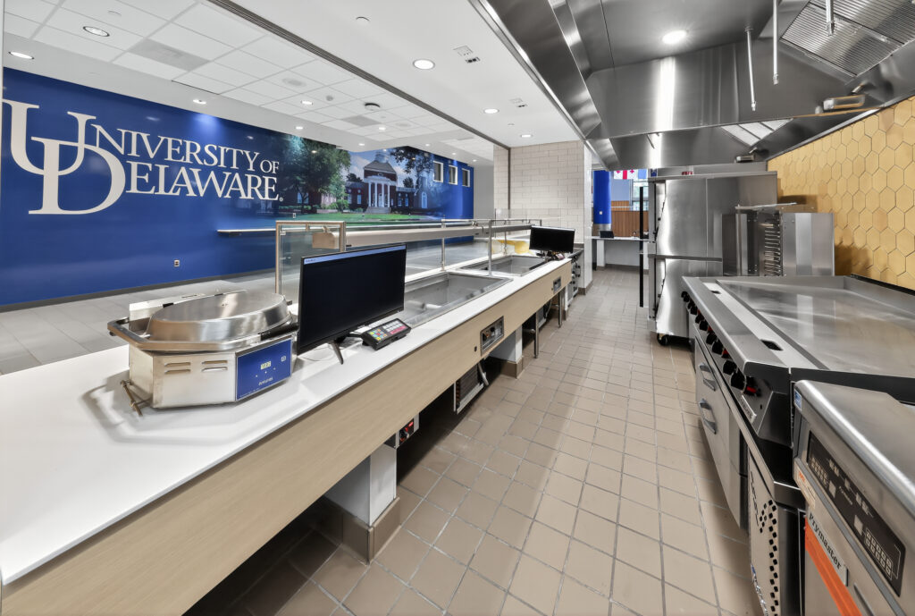 A commercial kitchen serving line with a University of Delaware logo on the wall and stainless steel appliances.