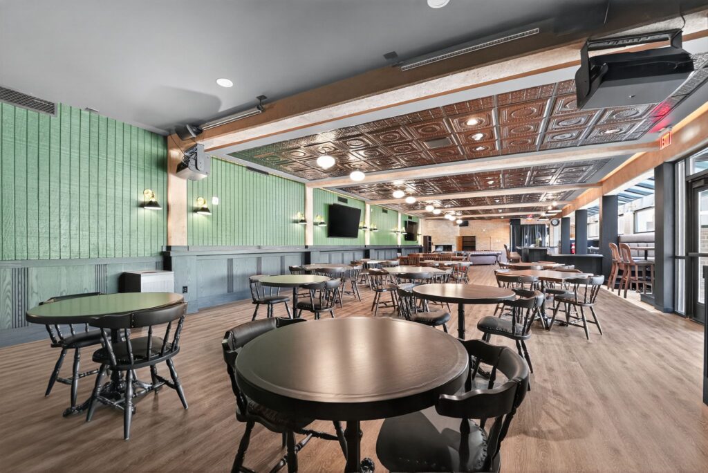 A restaurant dining room with green wood-paneled walls, a patterned metal ceiling, and several dark wood round tables with chairs.