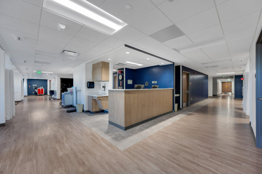 A hospital nursing station with a wood-fronted counter and blue accent wall in a wide hallway.