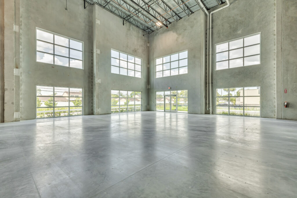 Empty warehouse interior with polished concrete floors, exposed ceiling beams, and large grid windows on three walls.