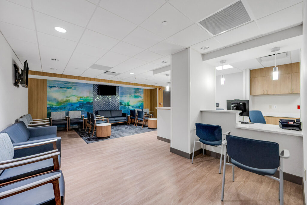 Waiting room with rows of blue chairs, a couch, a wall-mounted television, and a service desk.