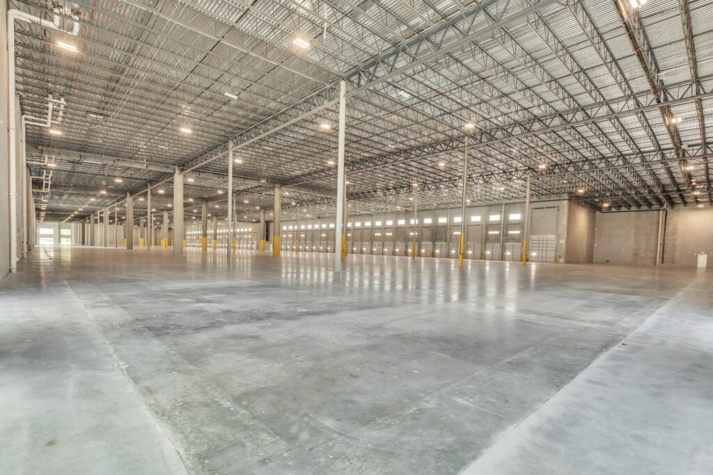 Large, empty warehouse interior with a concrete floor, high exposed metal ceiling, and a row of loading dock doors.