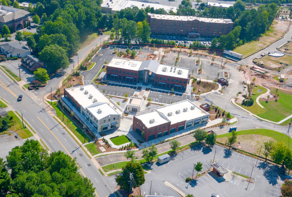 Aerial view of a commercial complex featuring several brick buildings with flat roofs, surrounding parking lots, and nearby wooded areas.