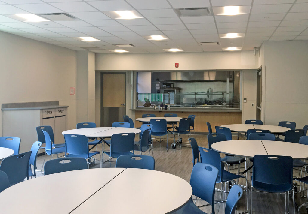 A school or office cafeteria with several round tables, blue chairs, and a kitchen service window in the back.