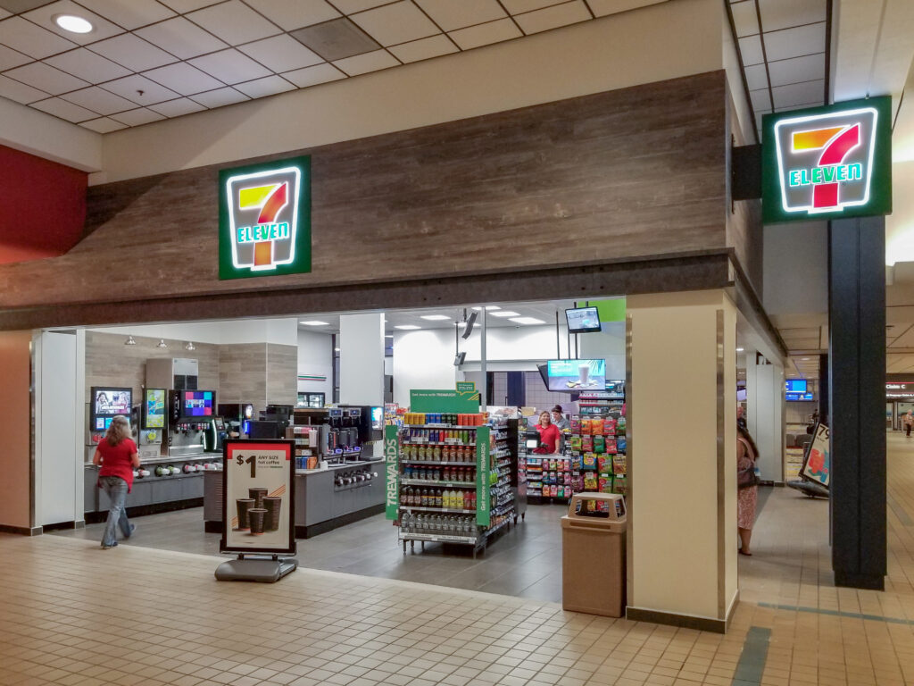 A 7-Eleven store entrance inside a building, showing drink dispensers, snack aisles, and a clerk behind the counter.