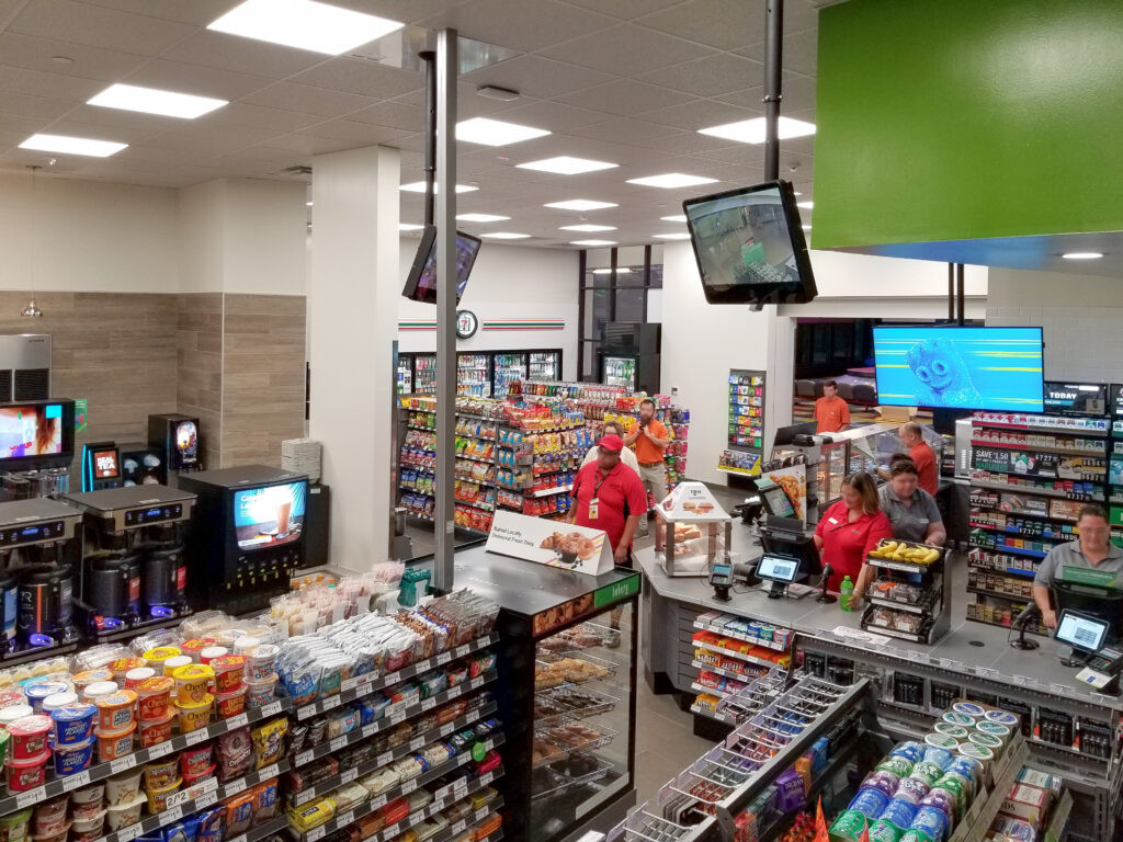 Interior of a 7-Eleven convenience store showing aisles of snacks, a coffee station, and employees working behind the checkout counter.