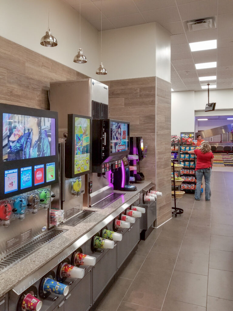 A row of slushy and soda fountain machines inside a convenience store, with a customer browsing nearby snack aisles.