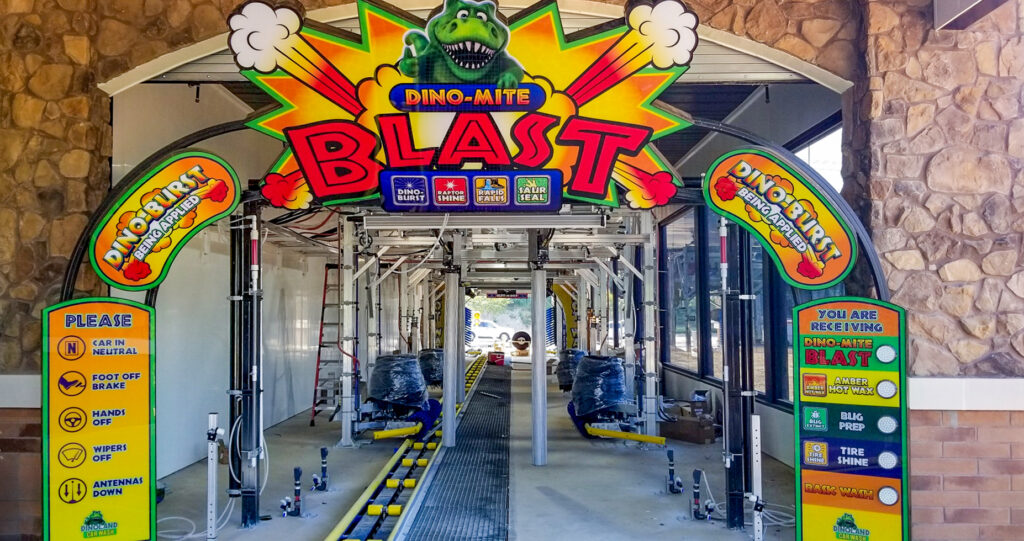 Entrance to a Dino-Lite car wash tunnel featuring large, colorful signage with instructions for drivers and lists of wash services.