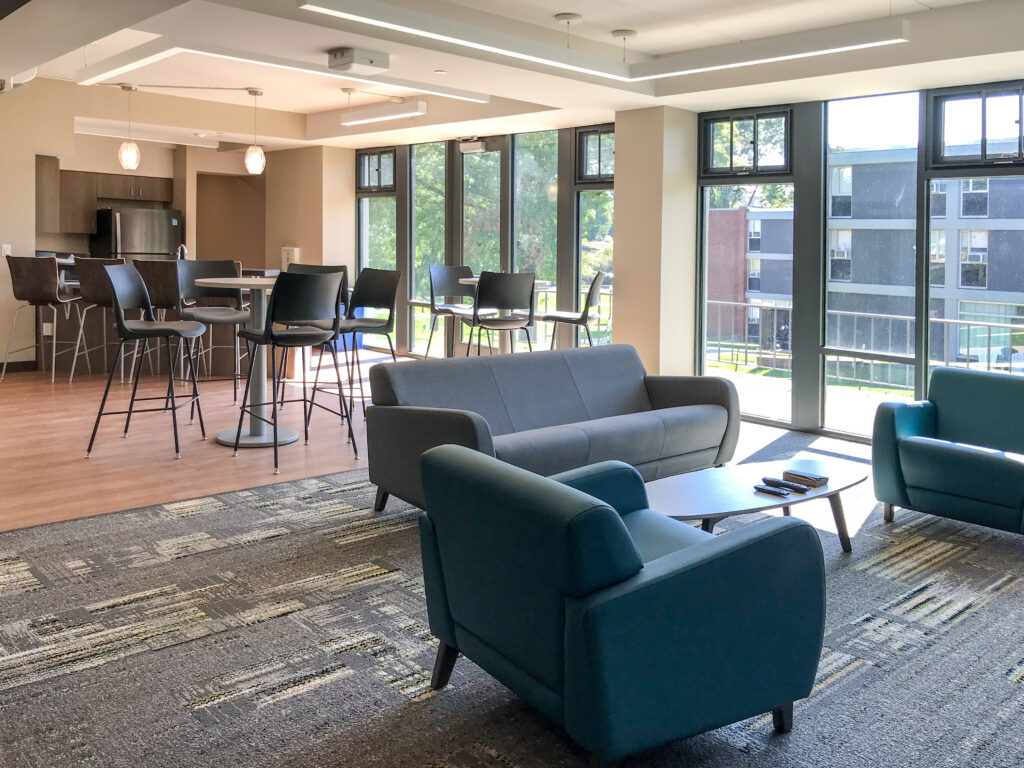A modern common area featuring a kitchenette with bar seating, a lounge space with a grey sofa and two blue armchairs, and floor-to-ceiling windows.