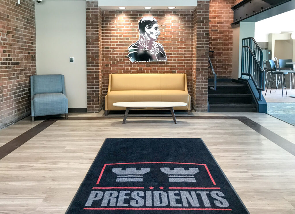 Lobby area with a cream-colored sofa, a coffee table, a blue armchair, and a floor mat featuring the word “PRESIDENTS” below a bust of Napoleon on a brick wall.