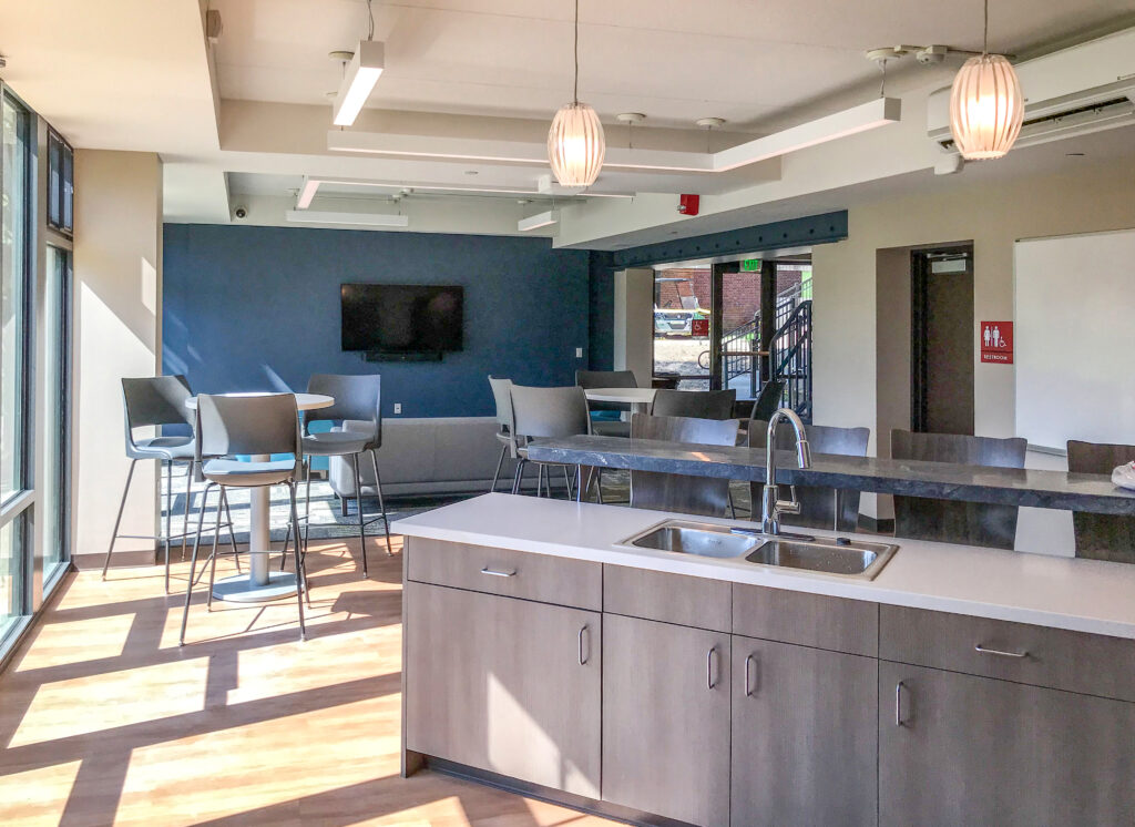 A communal kitchen and lounge area featuring a center island with a sink, small round tables with chairs, and a wall-mounted television.