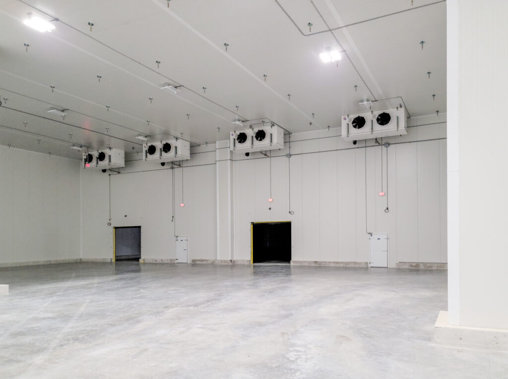 An empty, industrial cold storage warehouse with multiple ceiling-mounted cooling units and three doorways.