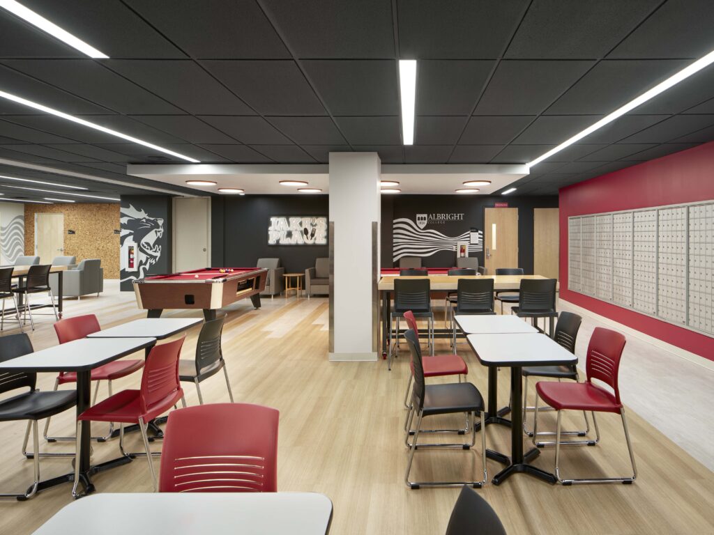 A brightly lit student lounge featuring red and black chairs, tables, a pool table, and a wall of mailboxes.