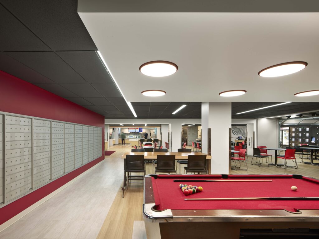 A pool table with red felt in a communal space featuring a wall of mailboxes, dining tables, and seating.