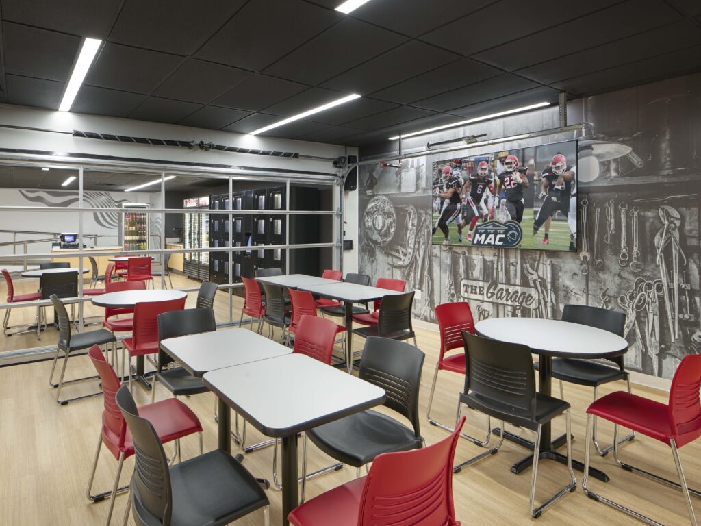 A cafeteria with white tables and black and red chairs, featuring a large screen showing a football game against a wall mural of mechanical tools.