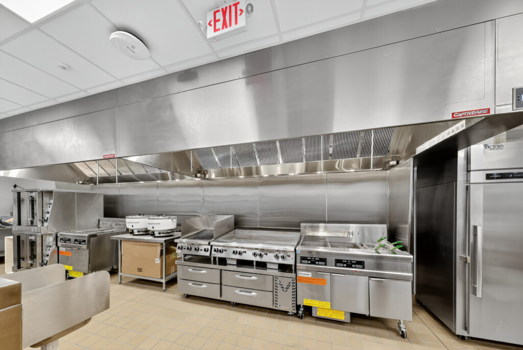 A commercial kitchen featuring stainless steel prep surfaces, a range with multiple burners, a flat-top grill, a deep fryer, and a large reach-in refrigerator under a ventilation hood.