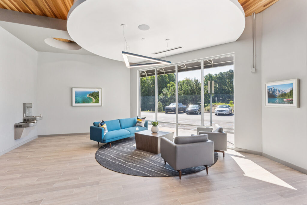 Waiting area with a blue sofa, two grey armchairs, a central coffee table, and large windows overlooking a parking lot.