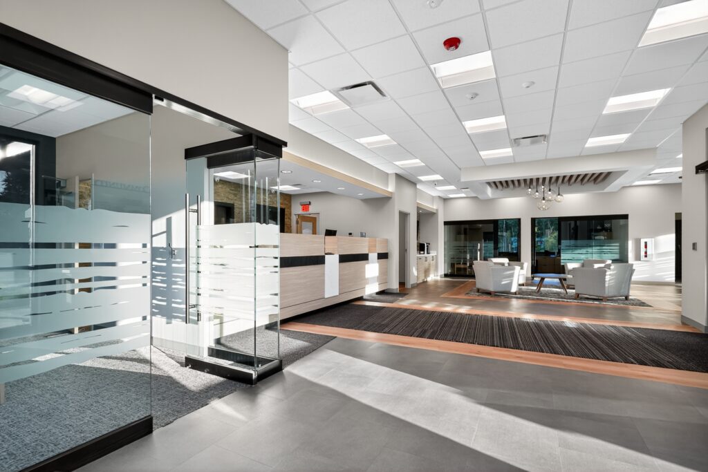 Spacious office lobby with a glass entryway, a long reception desk, and a seating area with chairs in the background.