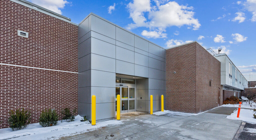 Exterior entrance of a brick building with a silver metal facade and yellow safety bollards, with patches of snow on the ground.