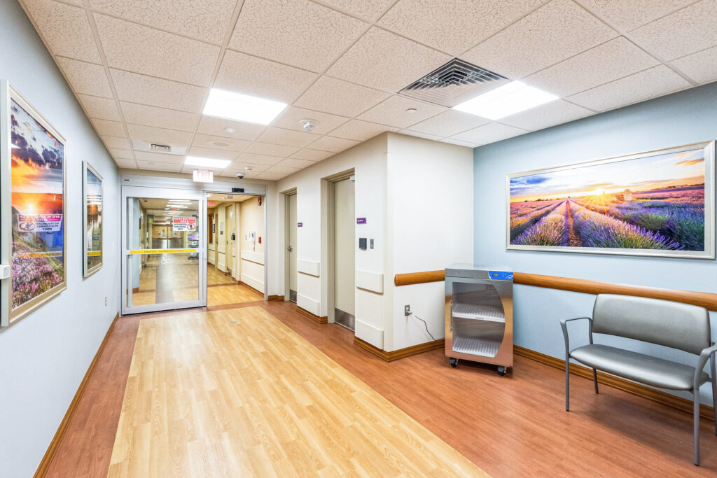 A hospital corridor with wood-look flooring, a sliding glass door entrance, a metal supply cart, and a waiting chair, decorated with landscape photographs on the walls.