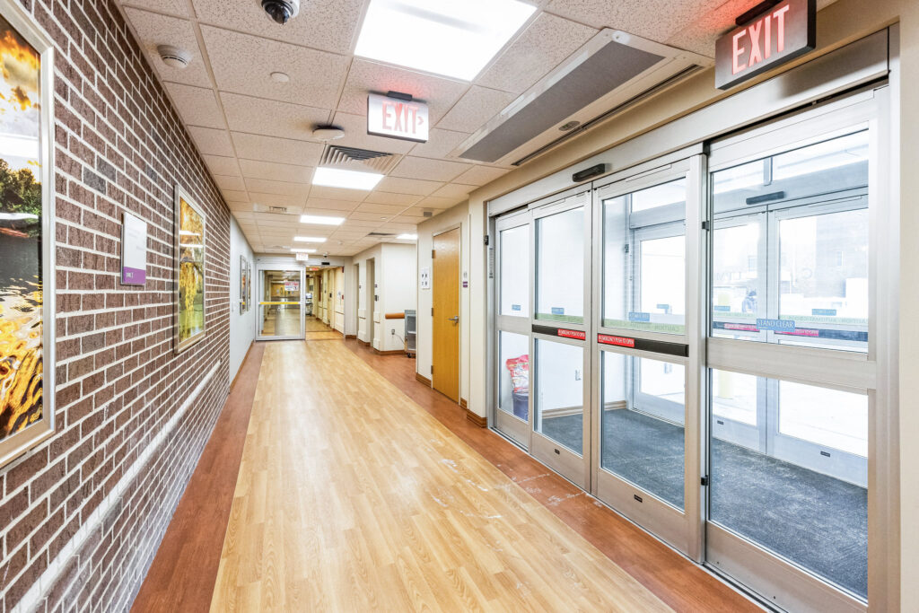 A long hospital corridor with wood-look flooring, brick walls, and automatic sliding glass exit doors.