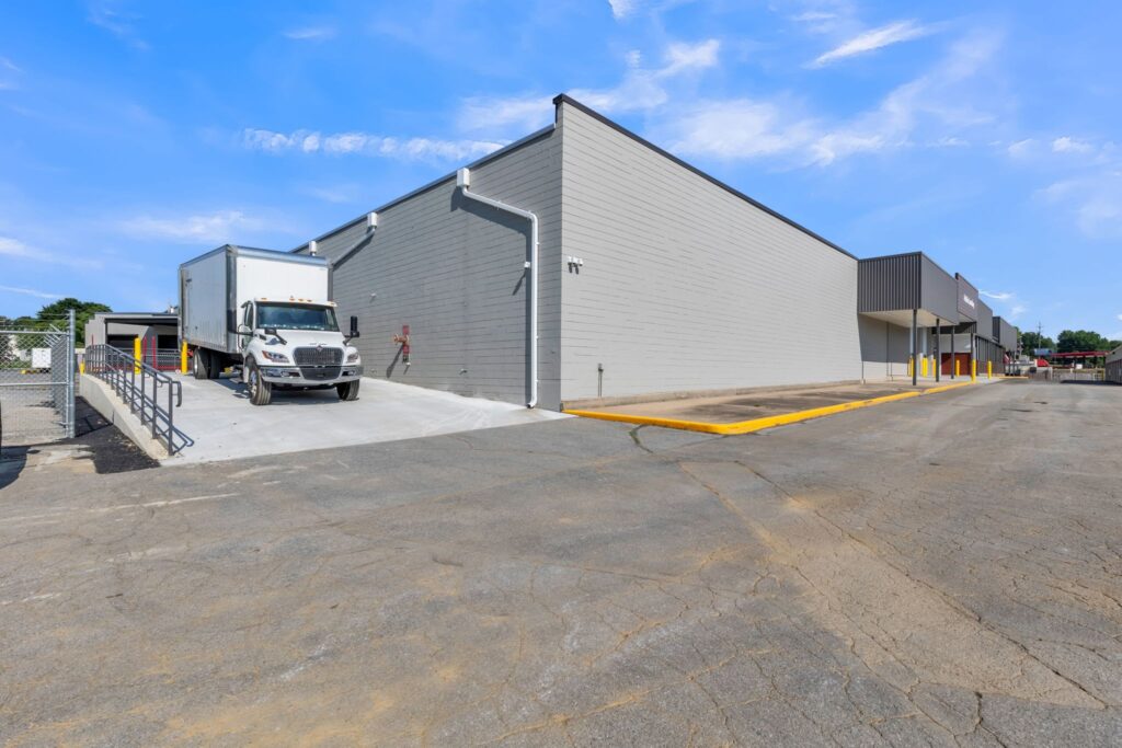 A semi-truck parked at the loading dock of a grey commercial warehouse building.