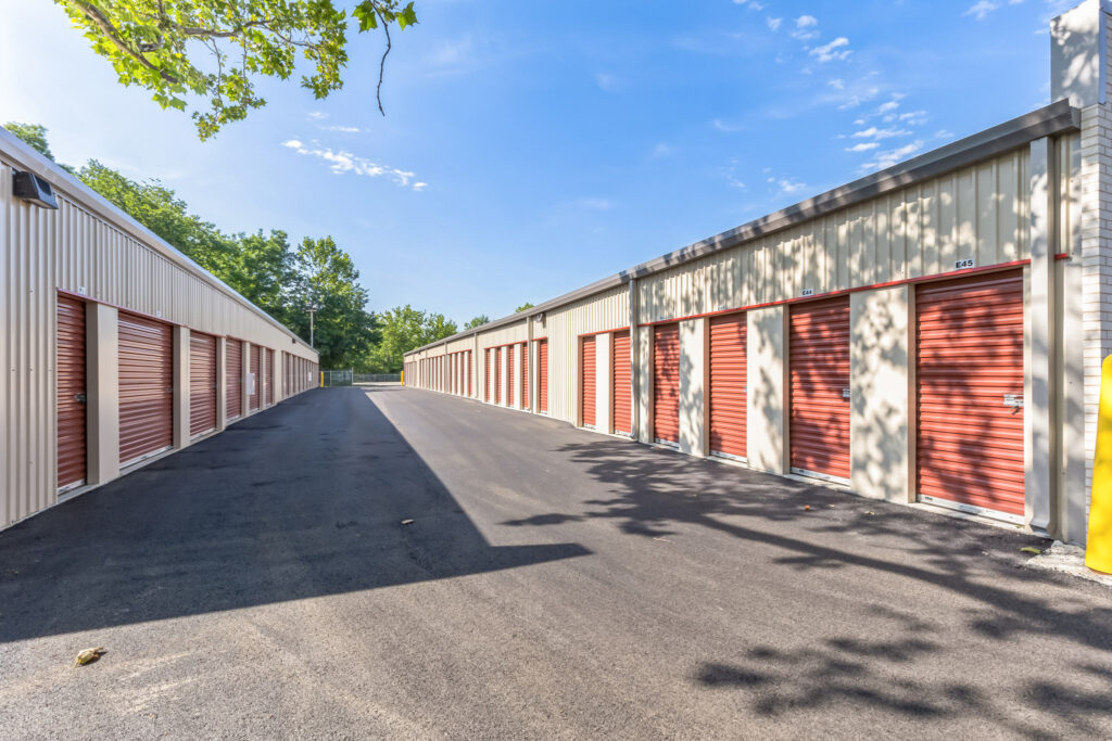 An outdoor self-storage facility with two rows of cream-colored buildings featuring red roll-up doors and unit numbers.