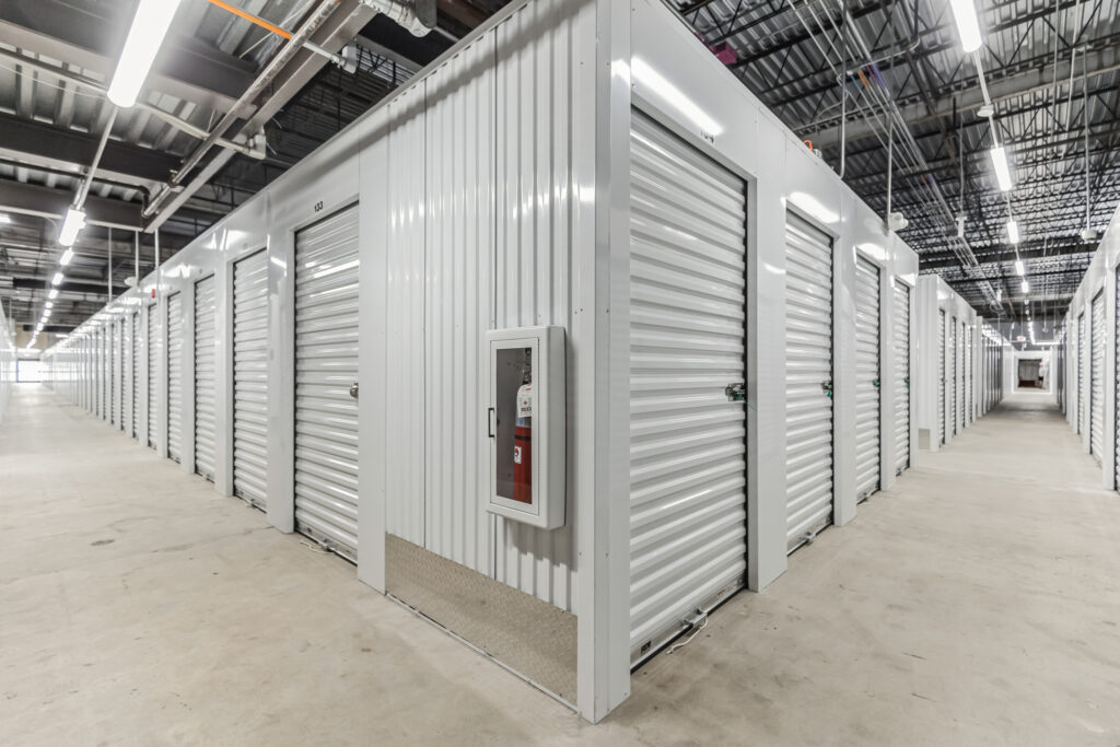 Rows of white metal storage units with rolling doors in an indoor facility, with a fire extinguisher mounted on the corner unit.