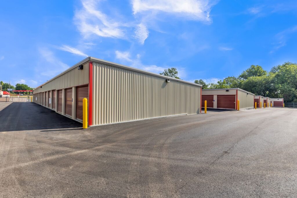 Exterior view of a self-storage facility with beige metal buildings and red roll-up doors, set against a bright blue sky.