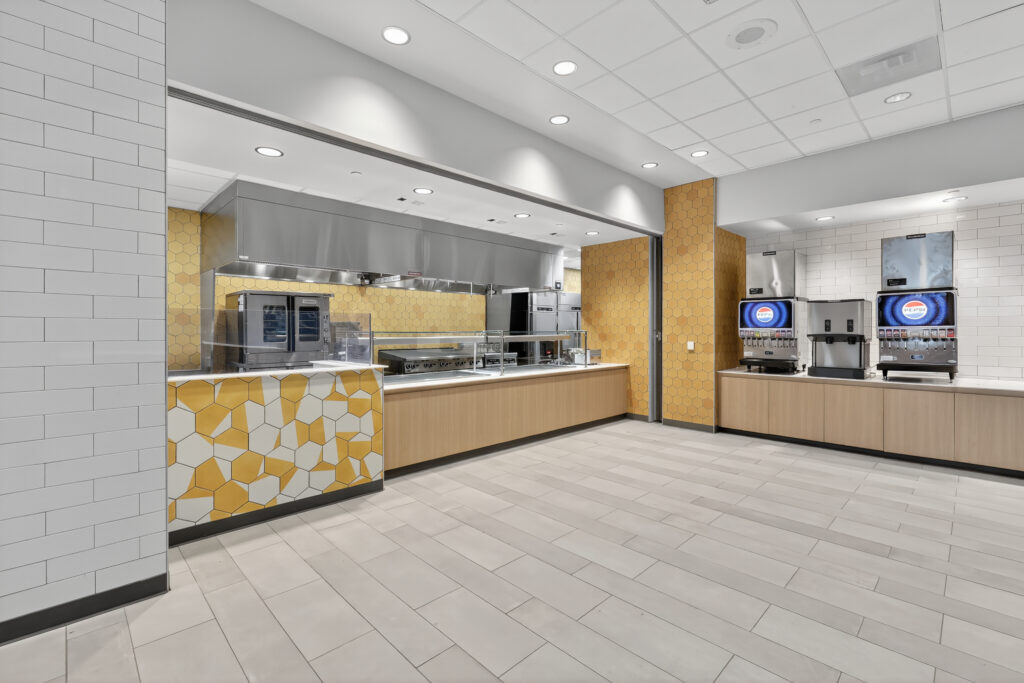 A modern commercial kitchen serving area with stainless steel ovens, a flat-top grill, and a row of beverage dispensers.