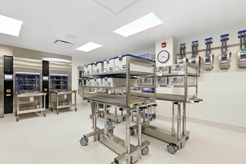 Two stainless steel carts holding plastic storage bins in a sterile hospital processing room, with stainless steel washing units in the background.