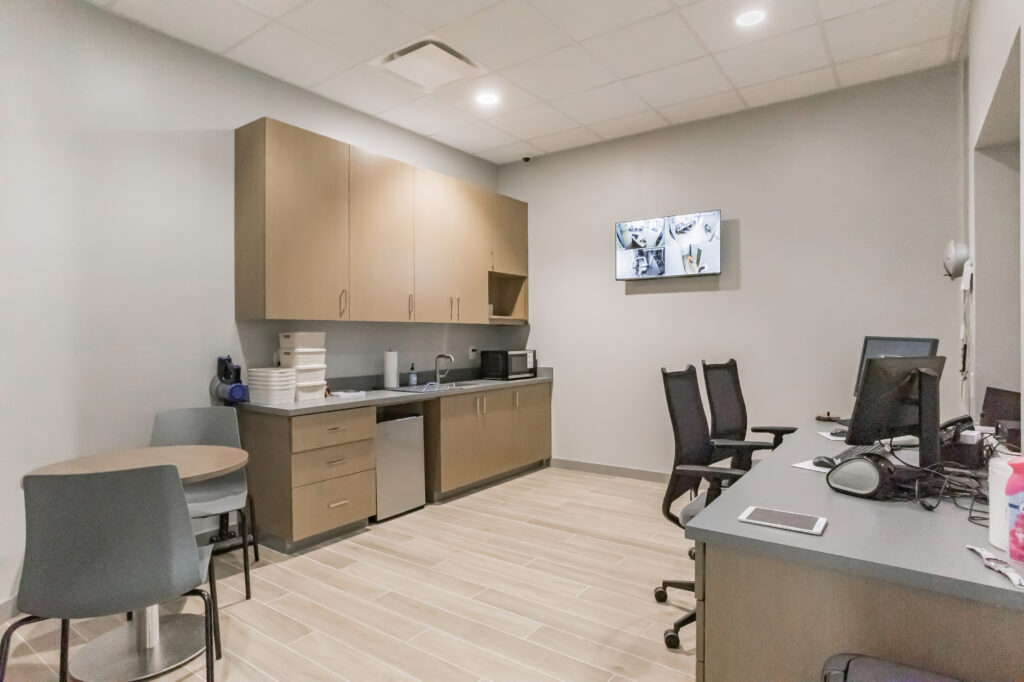 An office break room with a kitchenette, a small round table, and a desk with a computer monitor. A security camera display is mounted on the wall.
